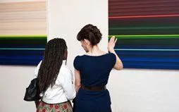 Photo of two ladies looking at an artwork on a wall. The picture is coloured bands of light stretching from left to right. The colours are mainly blues with some brighter red and green line. This was a giclee print on canvas stretched over wooden bearers.